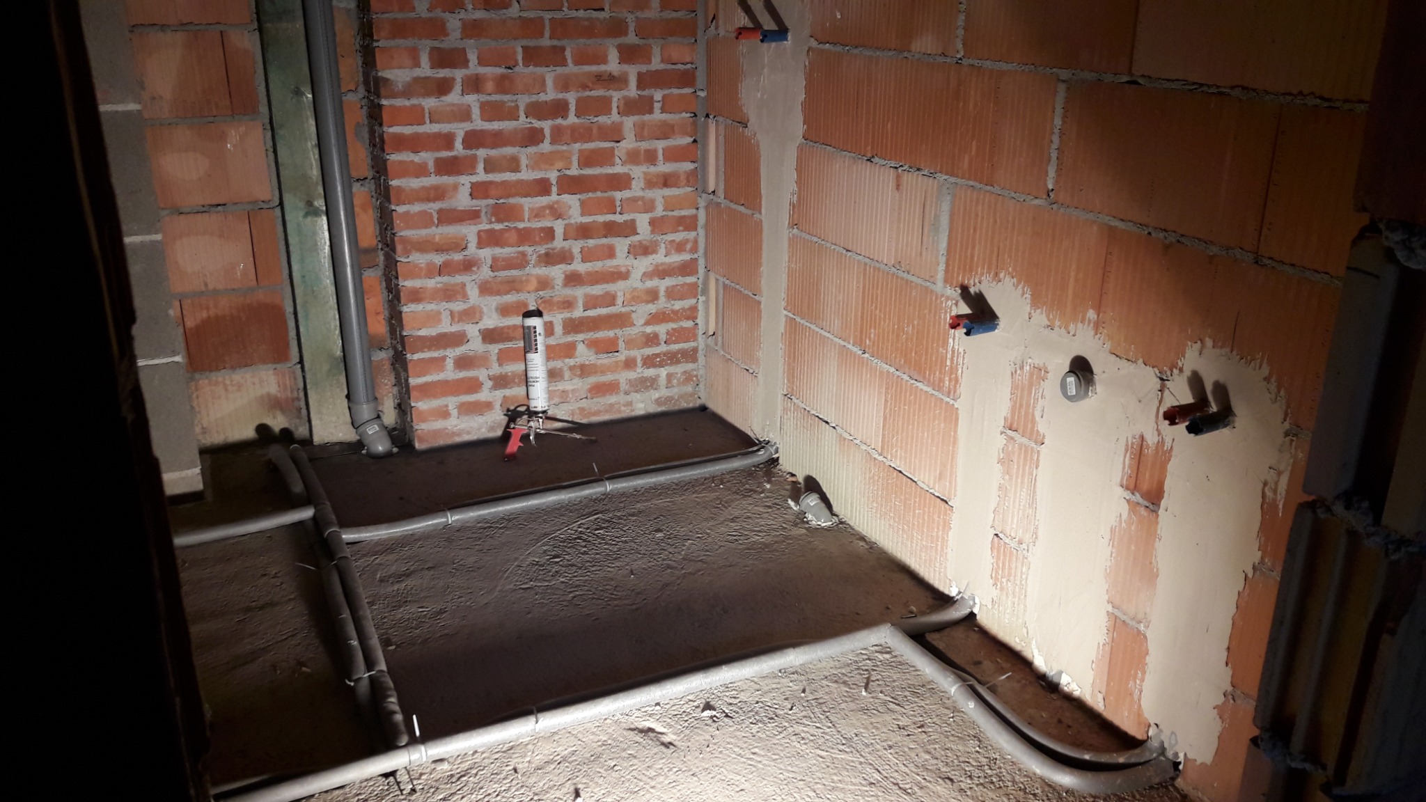 Surowy stan łazienki w trakcie prac: widoczne rury instalacji wodnej na podłodze, wystające przyłącza na ścianach z cegły dziurawki oraz pistolet do pianki montażowej.