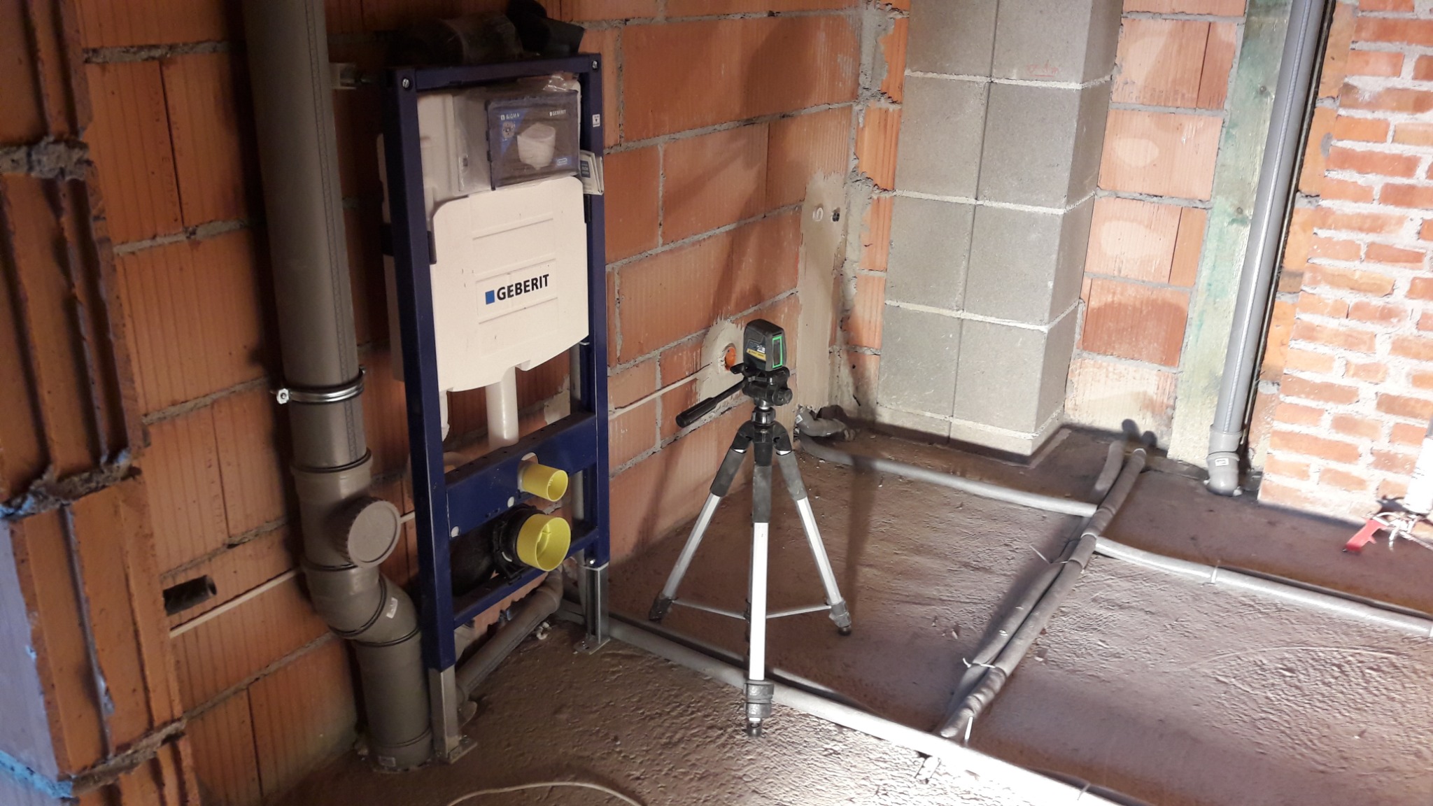 Stelaż podtynkowy Geberit zamontowany przy ścianie z cegły, z widocznymi rurami kanalizacyjnymi i poziomującym laserem na trójnogu, instalacja wodna w bruzdach w podłodze.