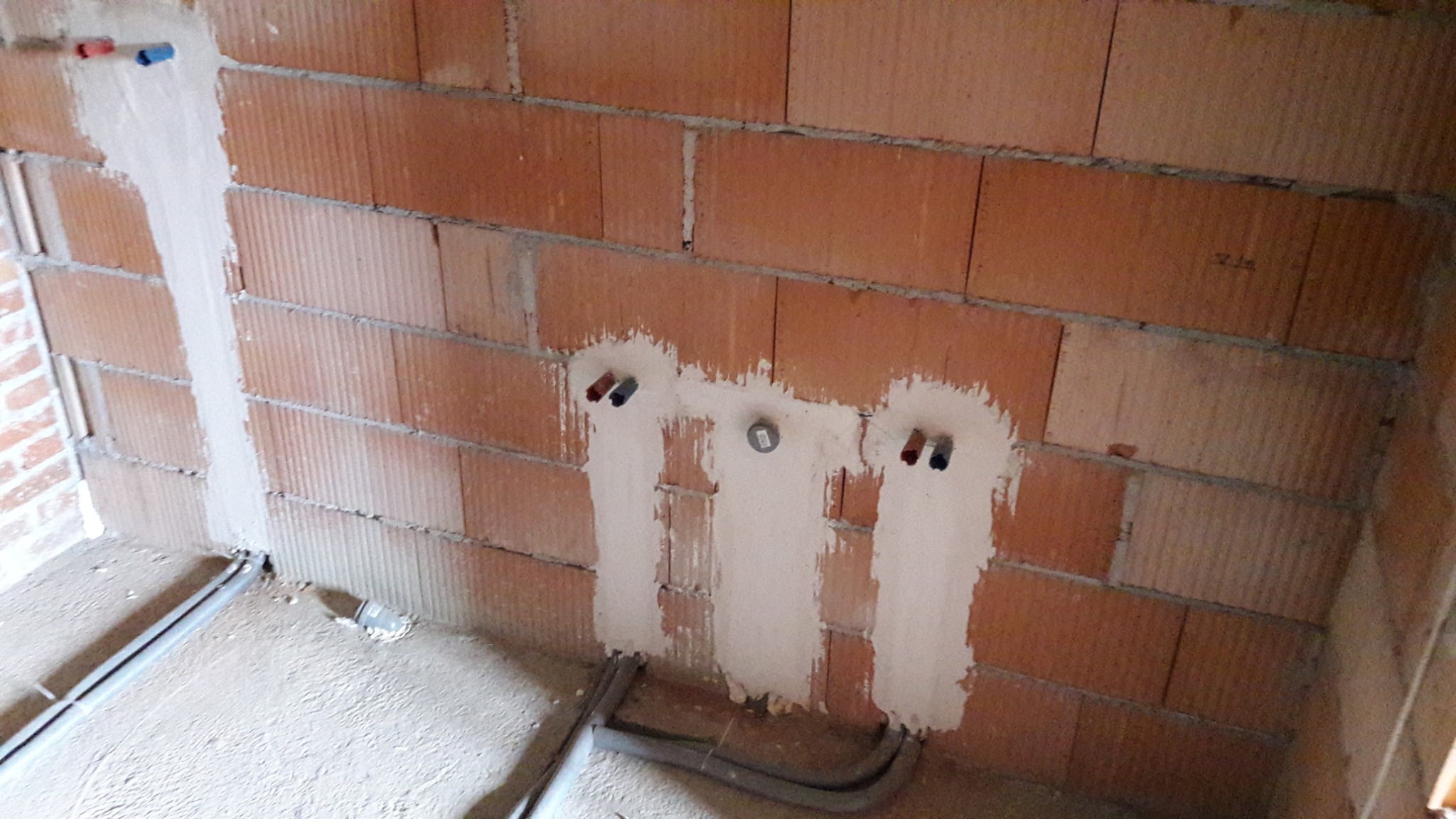 Ściana z czerwonej cegły z przygotowanymi wyprowadzeniami instalacji wodnej i grzewczej, zamurowane bruzdy z widocznymi rurami na podłodze, etap prac instalacyjnych w nowym budynku.