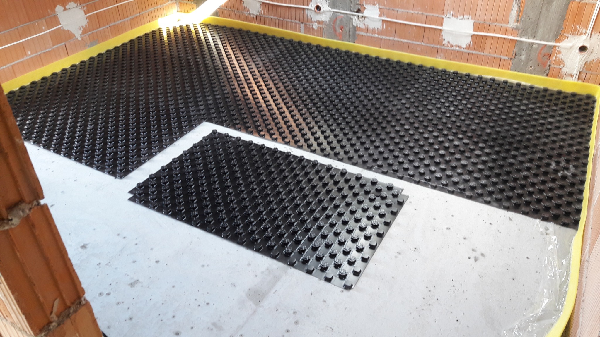 Czarna mata systemowa z wypustkami do montażu ogrzewania podłogowego, ułożona na folii ochronnej w surowym stanie, widoczne ściany z czerwonej cegły dziurawki.