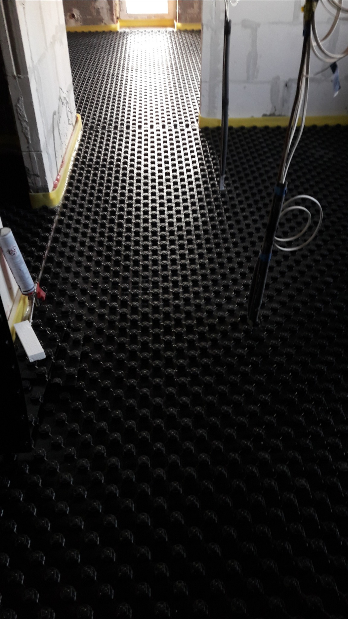 Czarna mata systemowa z wypustkami ułożona na podłodze w trakcie prac instalacyjnych, widoczne narzędzia i przewody, żółta taśma uszczelniająca przy ścianach.