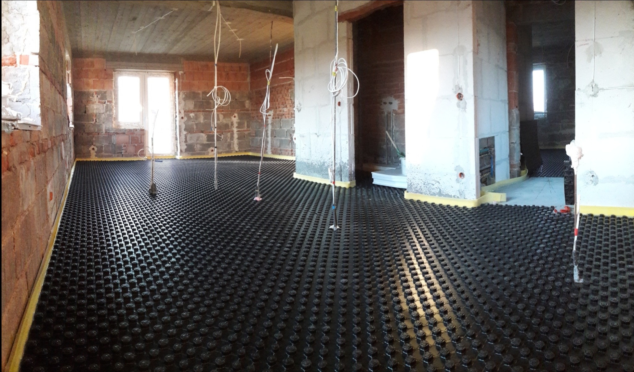 Rozległa instalacja ogrzewania podłogowego z czarną matą grzewczą pokrytą wypustkami w surowym stanie, widoczne przewody, ściany z surowej cegły i betonowy sufit w trakcie prac wykończeniowych.
