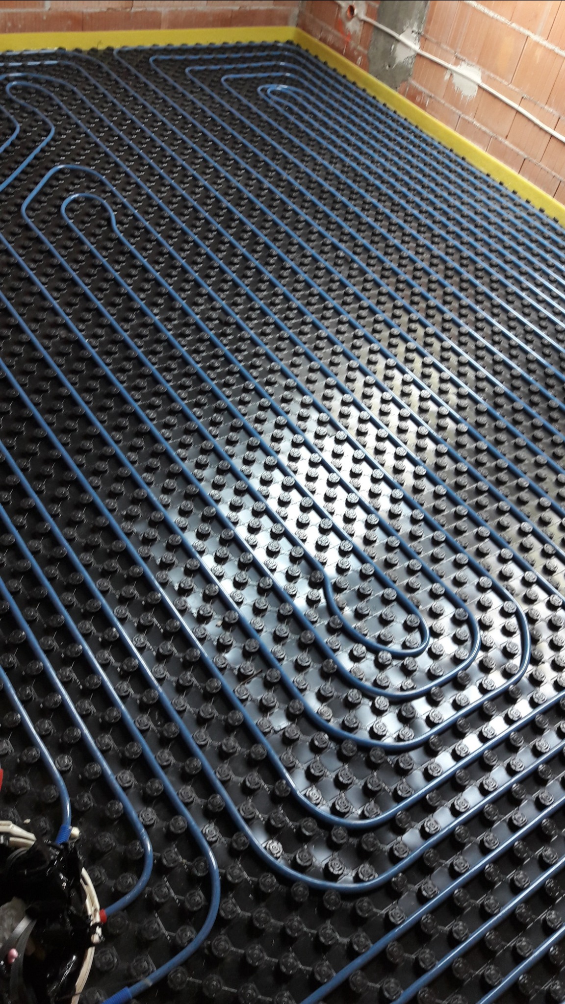 Niebieskie rury układane w pętle na czarnych uchwytach, tworzące system ogrzewania podłogowego na powierzchni z cegły i żółtą taśmą brzegową.
