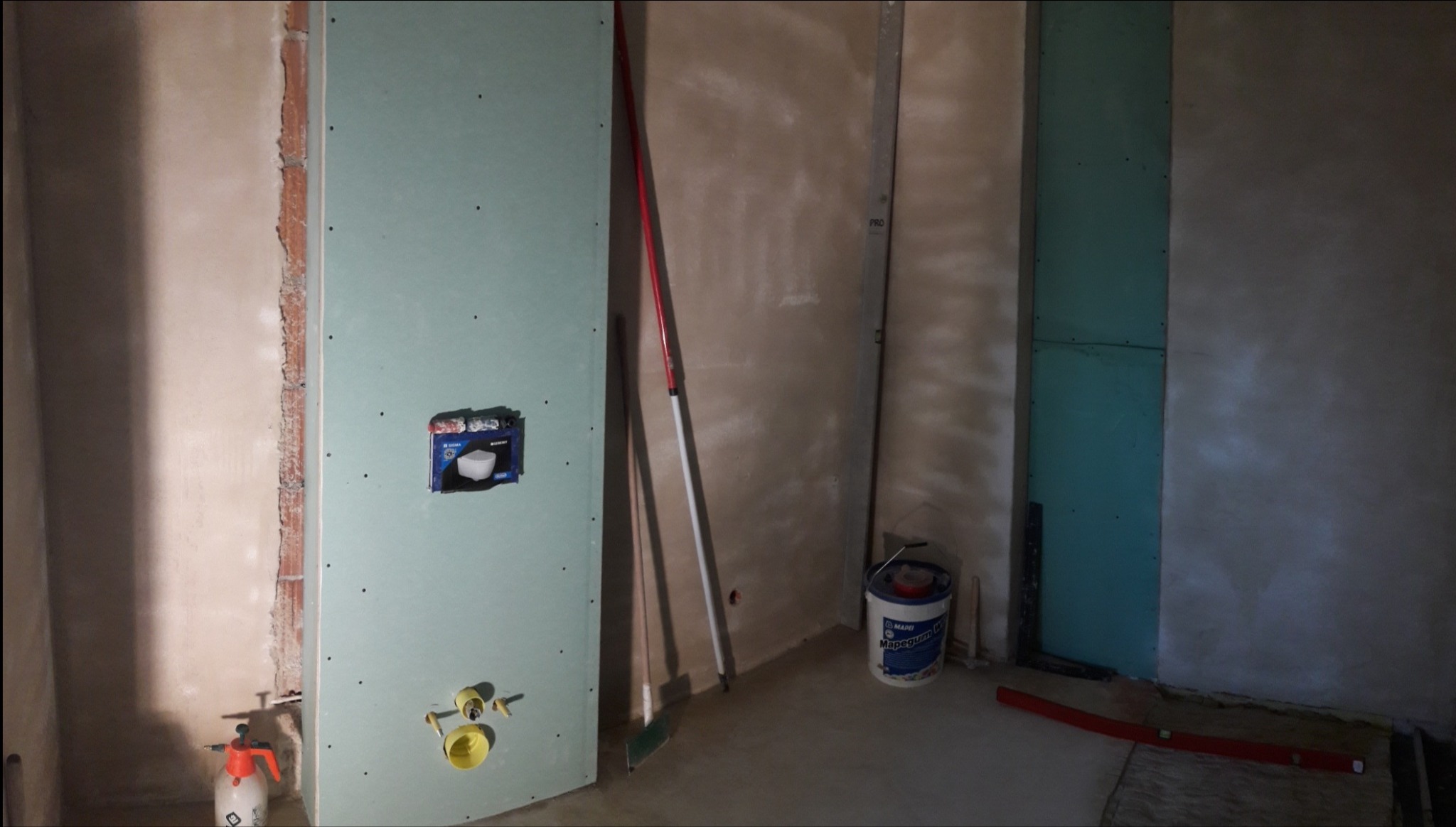 Wnętrze łazienki w trakcie prac wykończeniowych; widoczne płyty gipsowo-kartonowe na ścianach, elementy instalacji wodnej, narzędzia budowlane (poziomica, wiadro z zaprawą) oraz zamontowany stelaż...