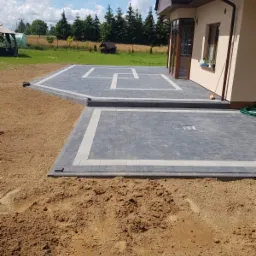 Nowo ułożona, szara kostka brukowa z białymi obramowaniami tworząca taras przy domu jednorodzinnym, widoczne przygotowane podłoże z piasku, w tle trawnik i drzewa.