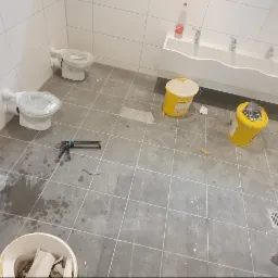 Wnętrze łazienki w przedszkolu podczas prac wykończeniowych: trzy toalety z nałożoną folią ochronną, rząd umywalek, wiaderka z farbą, narzędzia do fugowania i pozostałości po pracach na szarych...