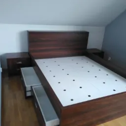 Łóżko z ciemnego drewna z wysuwanymi szufladami i białym stelażem z otworami, dopasowane szafki nocne, ustawione na drewnianej podłodze w pokoju z białymi i szarymi ścianami.