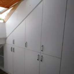 Biała szafa na wymiar zabudowana pod skosem dachu, z metalowymi uchwytami i drewnianym żaluzjami w oknie dachowym.