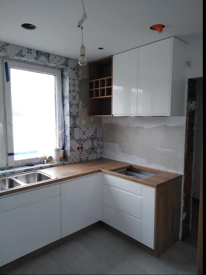 Wnętrze kuchni w trakcie montażu: białe, lakierowane szafki wiszące z otwartą półką na wino, drewniany blat, białe szafki dolne z szufladami, płytki dekoracyjne na ścianie, widoczne niedokończone...