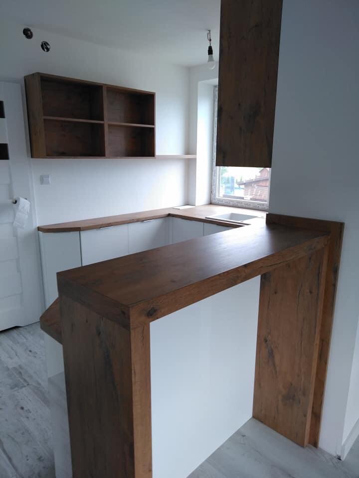 Jasna kuchnia z drewnianym blatem i barkiem, widoczne półki wiszące i okno z widokiem na zabudowę.