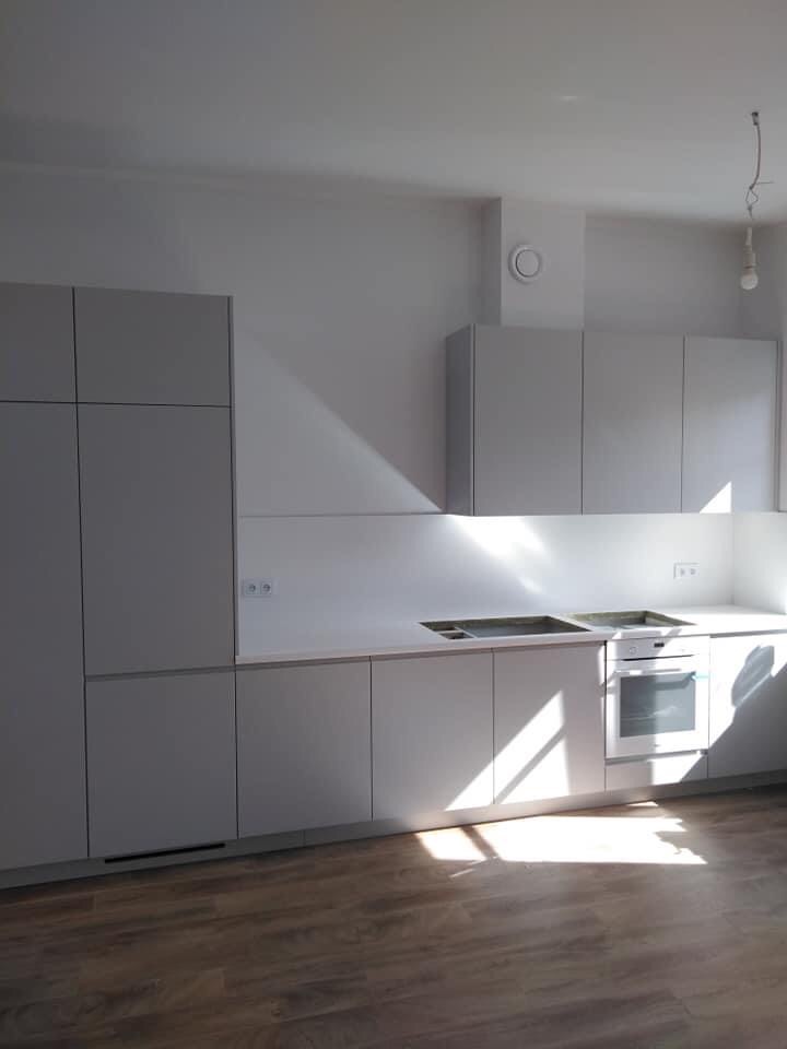 Nowoczesna kuchnia z szarymi, matowymi frontami szafek, jasnym blatem i drewnopodobną podłogą, oświetlona silnym, naturalnym światłem słonecznym wpadającym przez okno.