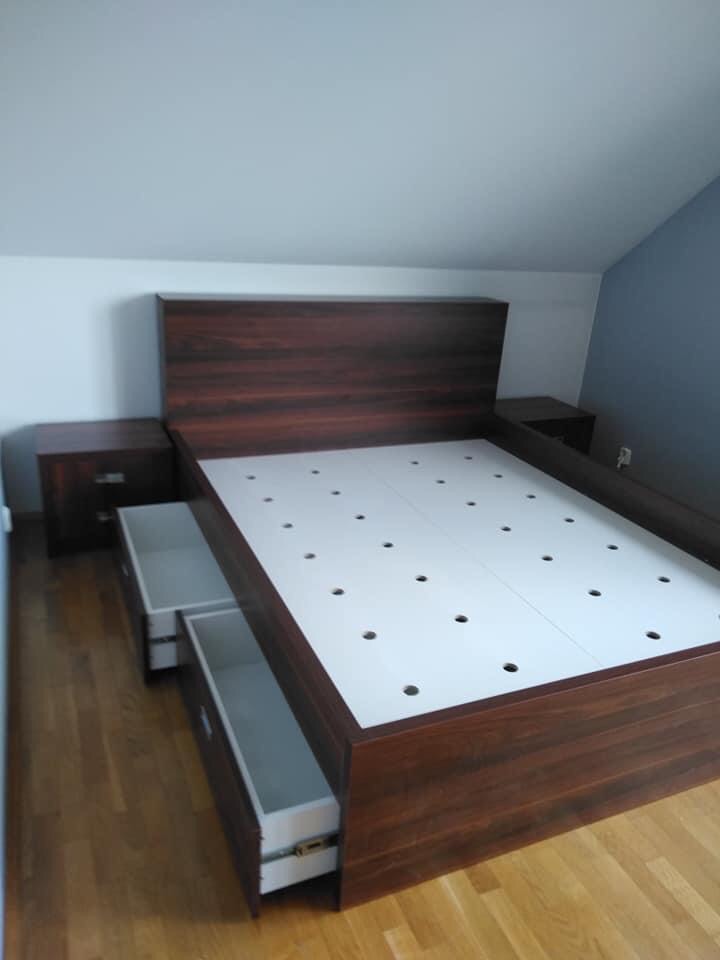 Łóżko z ciemnego drewna z wysuwanymi szufladami i białym stelażem z otworami, dopasowane szafki nocne, ustawione na drewnianej podłodze w pokoju z białymi i szarymi ścianami.