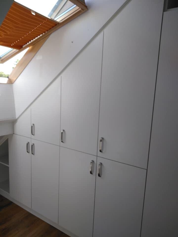 Biała szafa na wymiar zabudowana pod skosem dachu, z metalowymi uchwytami i drewnianym żaluzjami w oknie dachowym.