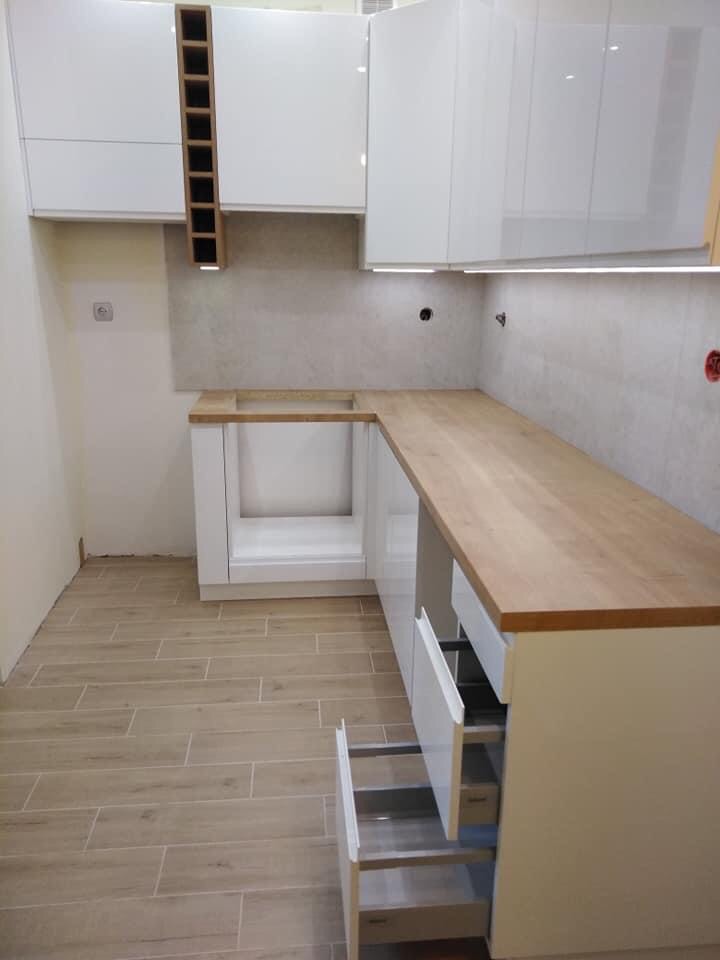 Nowoczesna, biała zabudowa kuchenna z drewnianym blatem i otwartymi szufladami, widoczne płytki imitujące drewno na podłodze.