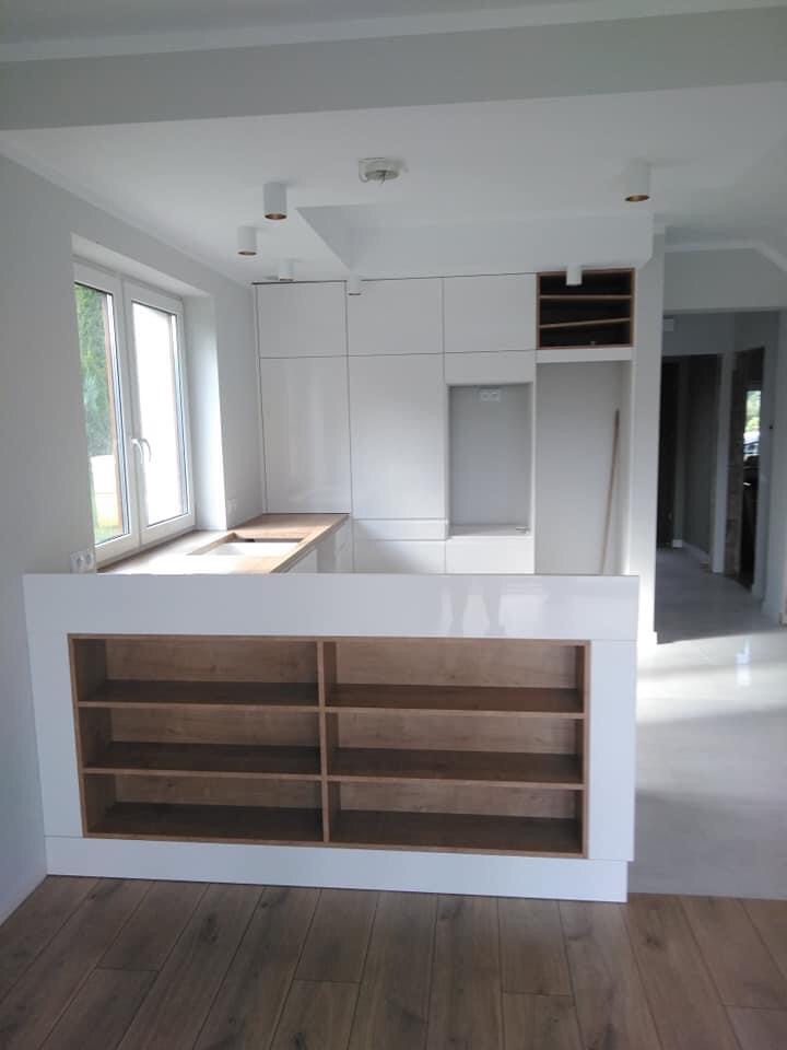 Nowoczesna, biała kuchnia z wyspą z otwartymi półkami z jasnego drewna, widoczna w trakcie montażu, z niekompletnym blatem i surowym wykończeniem wnętrza.