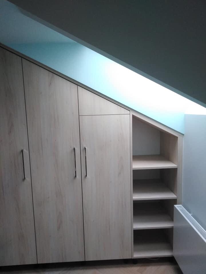 Szafa na wymiar z jasnego drewna, dopasowana do skosu poddasza, z częścią zamykaną na drzwi i otwartymi półkami po prawej stronie, obok grzejnik.