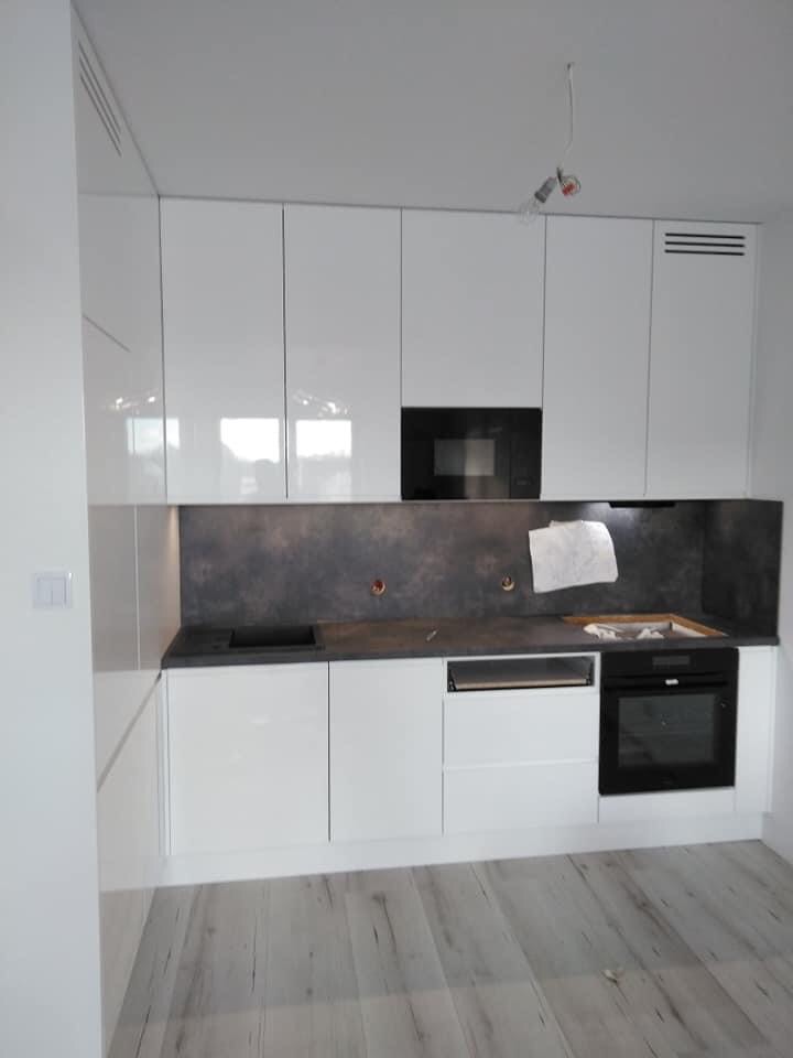 Nowoczesna, biała zabudowa kuchenna z czarnymi akcentami, szarym blatem i backsplashiem, widok na fragment podłogi z paneli imitujących drewno, surowa żarówka zwisająca z sufitu.
