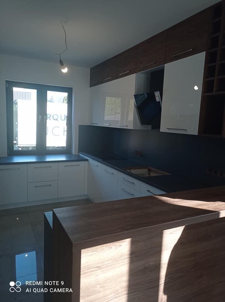 Nowoczesna kuchnia z białymi frontami szafek, ciemnym blatem i drewnianym barkiem. Górne szafki w kolorze drewna i białe, zintegrowany okap. Widoczne okno z czarną ramą i wisząca lampa.