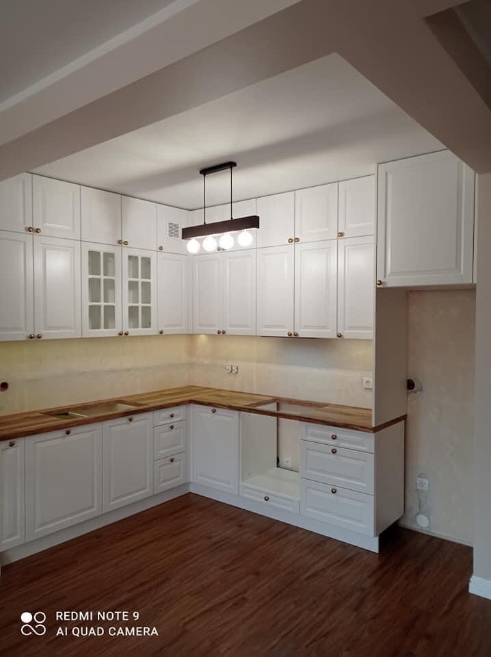 Jasna kuchnia z białymi szafkami i drewnianymi blatami, oświetlona podłużną lampą z czterema żarówkami. Widoczny fragment ściany z niedokończonym wykończeniem.