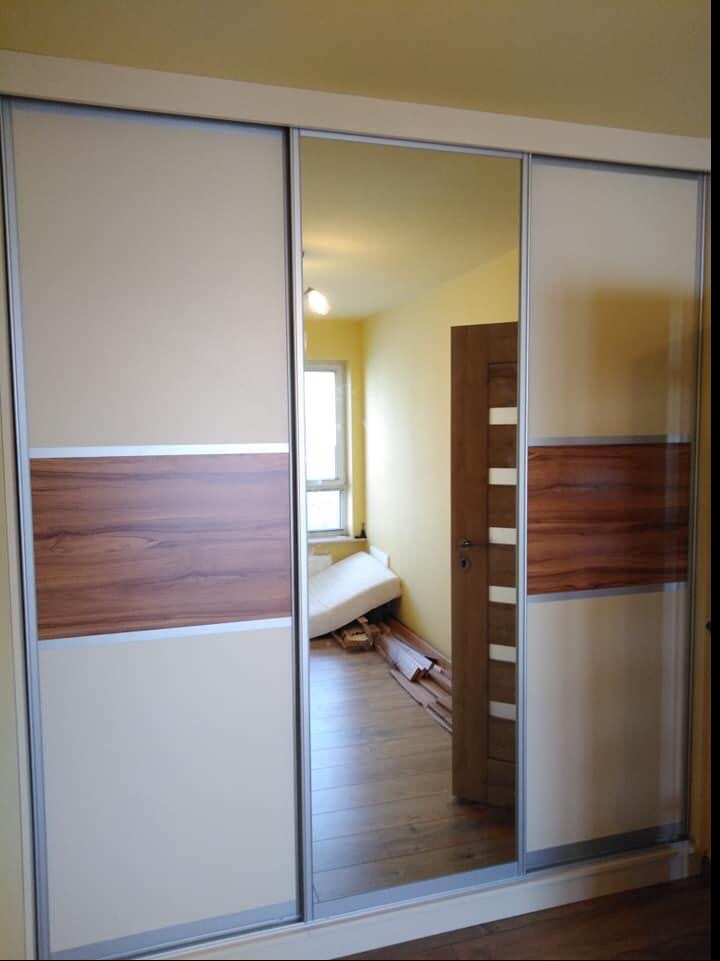 Trzydrzwiowa szafa przesuwna z lustrzanym środkowym panelem, beżowymi i drewnopodobnymi wstawkami, odbijająca wnętrze pokoju z rozłożoną sofą i niedokończonymi pracami wykończeniowymi.