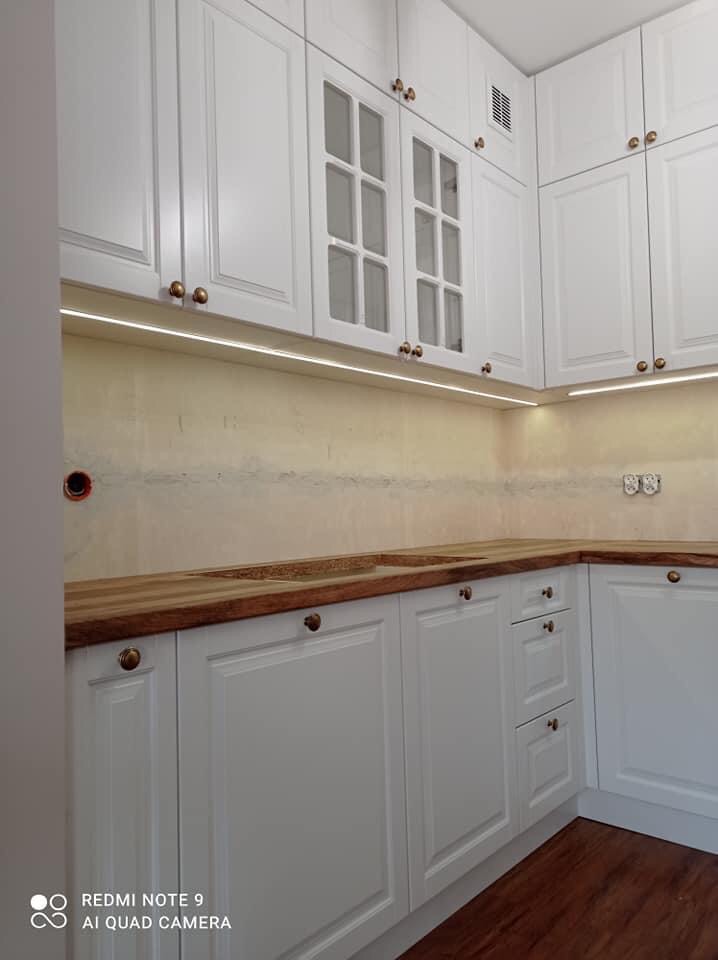 Jasna kuchnia z białymi szafkami w stylu klasycznym, drewnianym blatem i oświetleniem LED podszafkowym. Widoczne przeszklone szafki wiszące i mosiężne uchwyty.