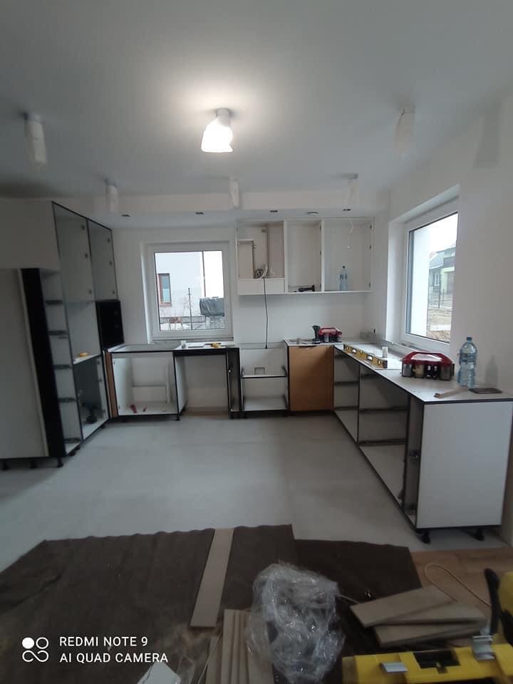 Montaż białych szafek kuchennych z czarnymi elementami, widoczne narzędzia i materiały budowlane na podłodze, okna w tle, surowy stan wykończenia wnętrza.