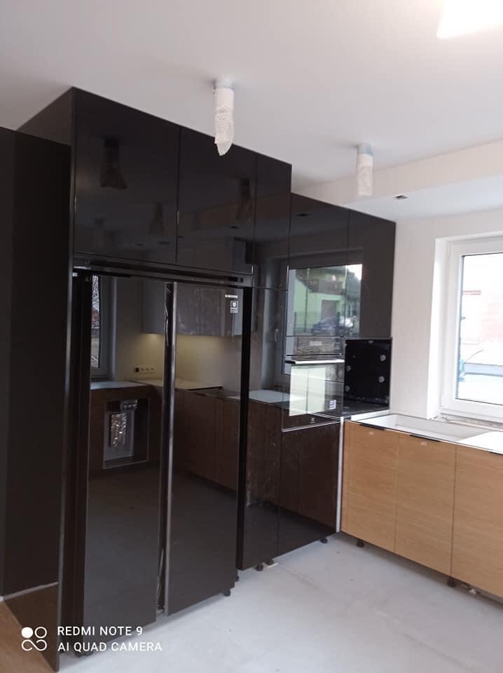 Nowoczesna kuchnia z czarnymi, lakierowanymi szafkami górnymi i drewnianymi szafkami dolnymi, widoczna lodówka side-by-side oraz zabudowany piekarnik, lampy sufitowe zabezpieczone folią ochronną.