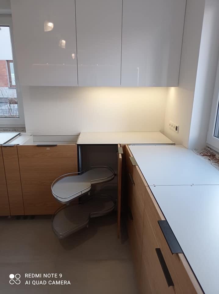 Nowoczesna kuchnia z jasnego drewna i białymi, błyszczącymi szafkami górnymi, prezentująca otwarty narożny system półek obrotowych.