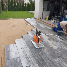 Montaż szarej kostki brukowej na podsypce piaskowej przy użyciu zagęszczarki w pobliżu garażu i trawnika z tujami.