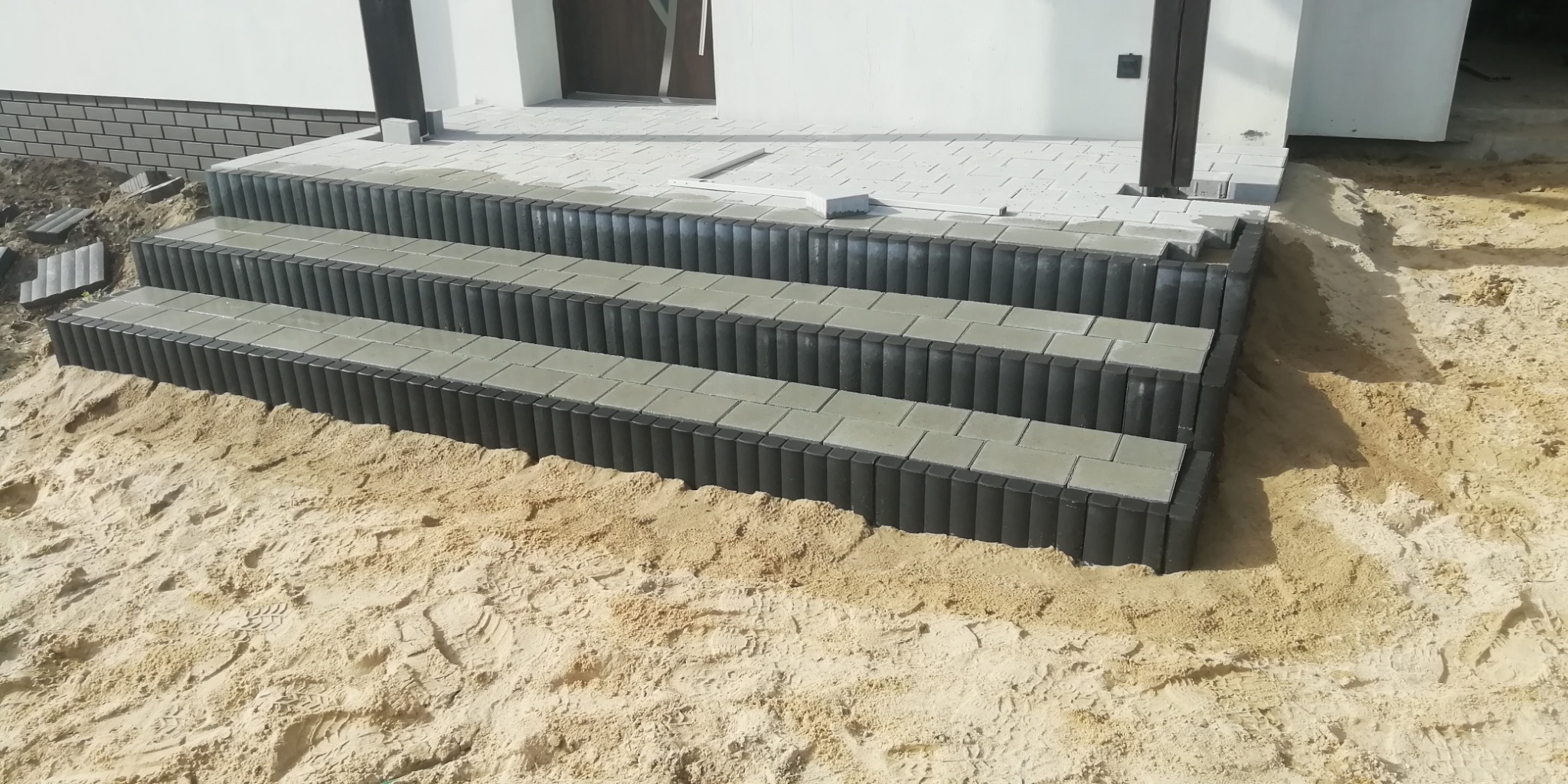 Trzy stopnie schodów wejściowych wykończone szarą kostką brukową i obrzeżami z ciemnych, pionowych elementów, otoczone piaskiem.