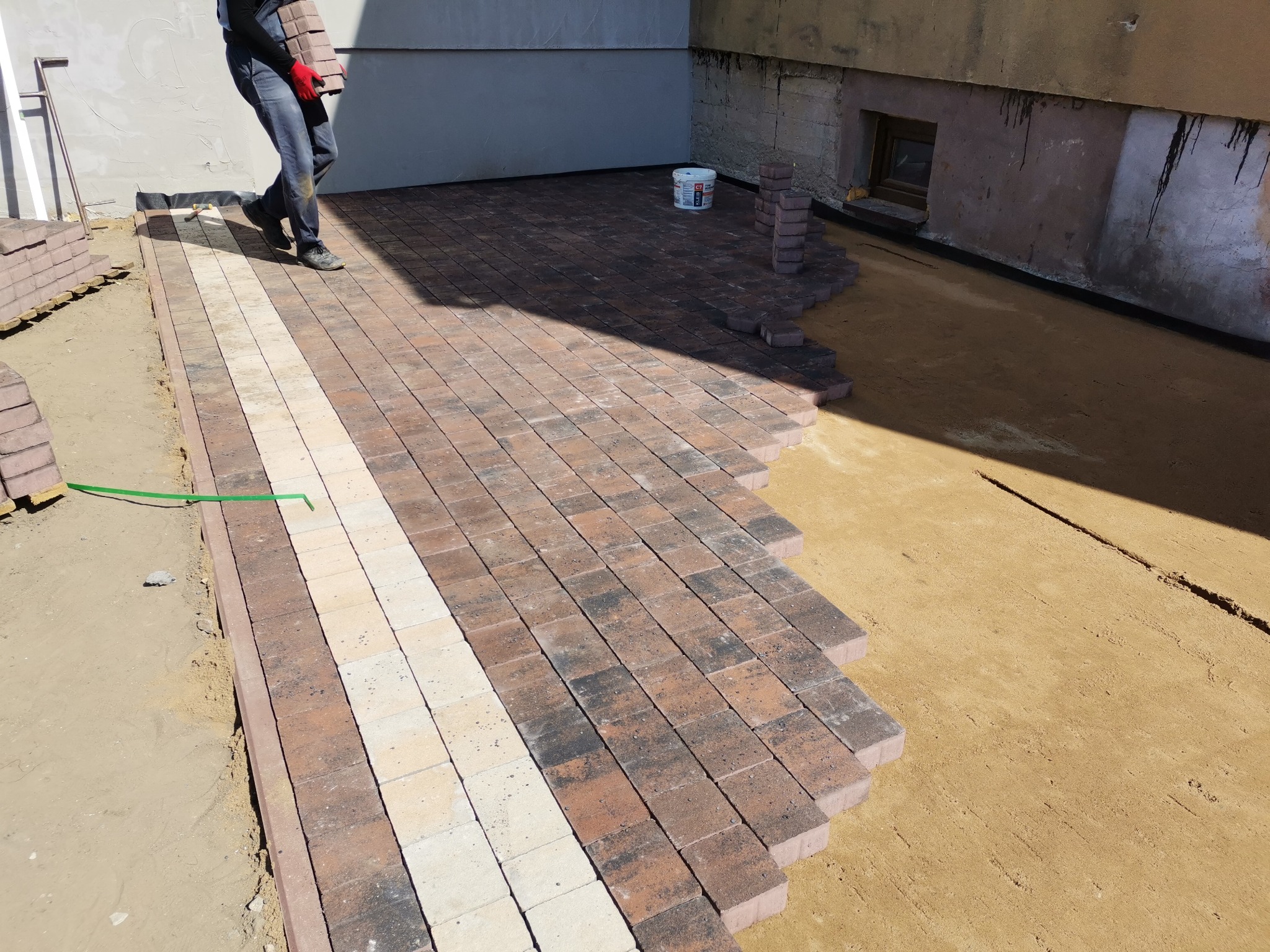 Prace brukarskie w toku: układanie wielobarwnej kostki na podsypce piaskowej, widoczny fragment ułożonej nawierzchni z jasnym pasem, pracownik w oddali z kostką, wiadro z klejem i stos kostki przy...