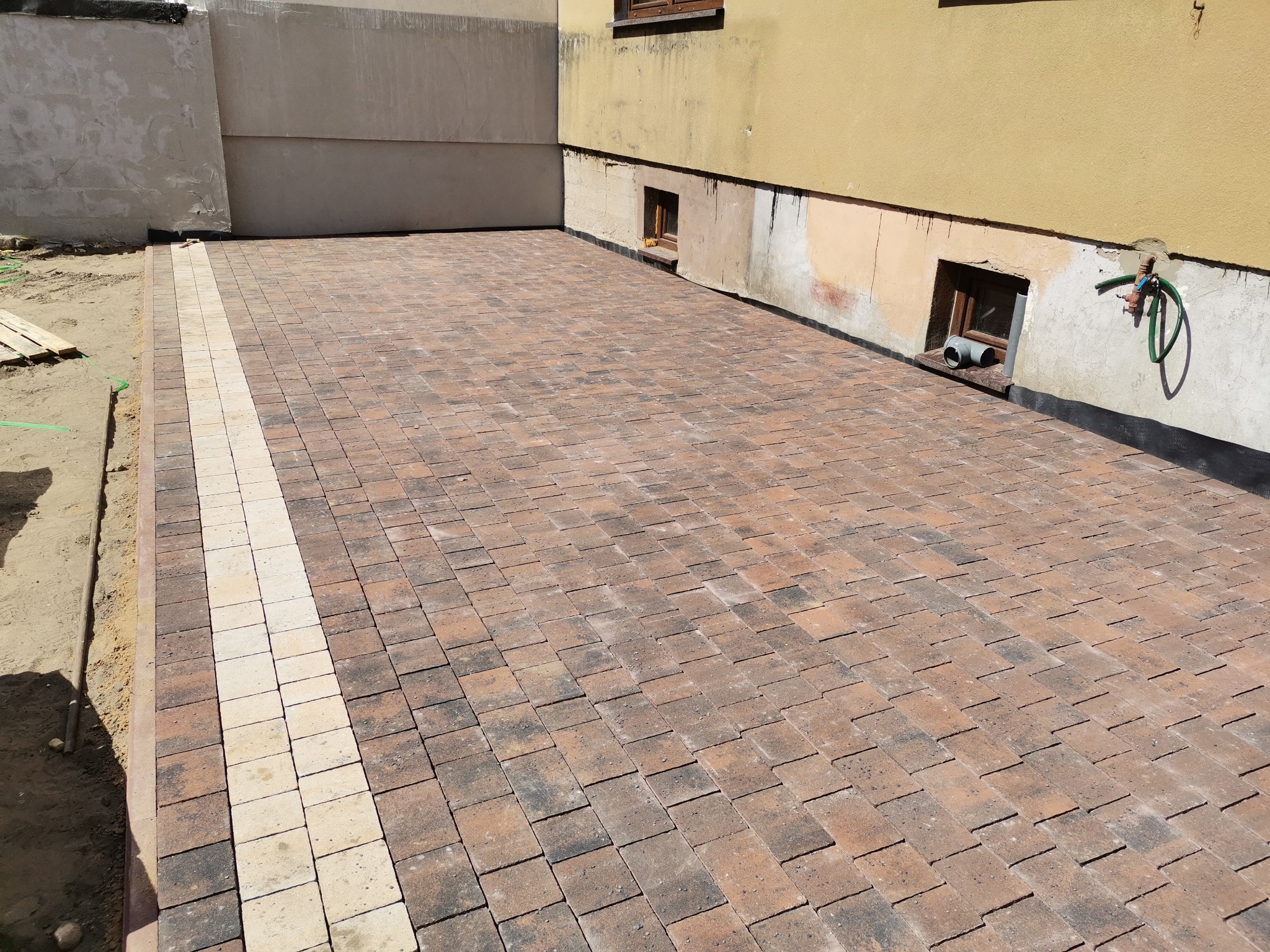 Ułożona kostka brukowa w kolorze brązowo-czerwonym z jasnym pasem na obrzeżach posesji, widoczne fragmenty elewacji budynku.