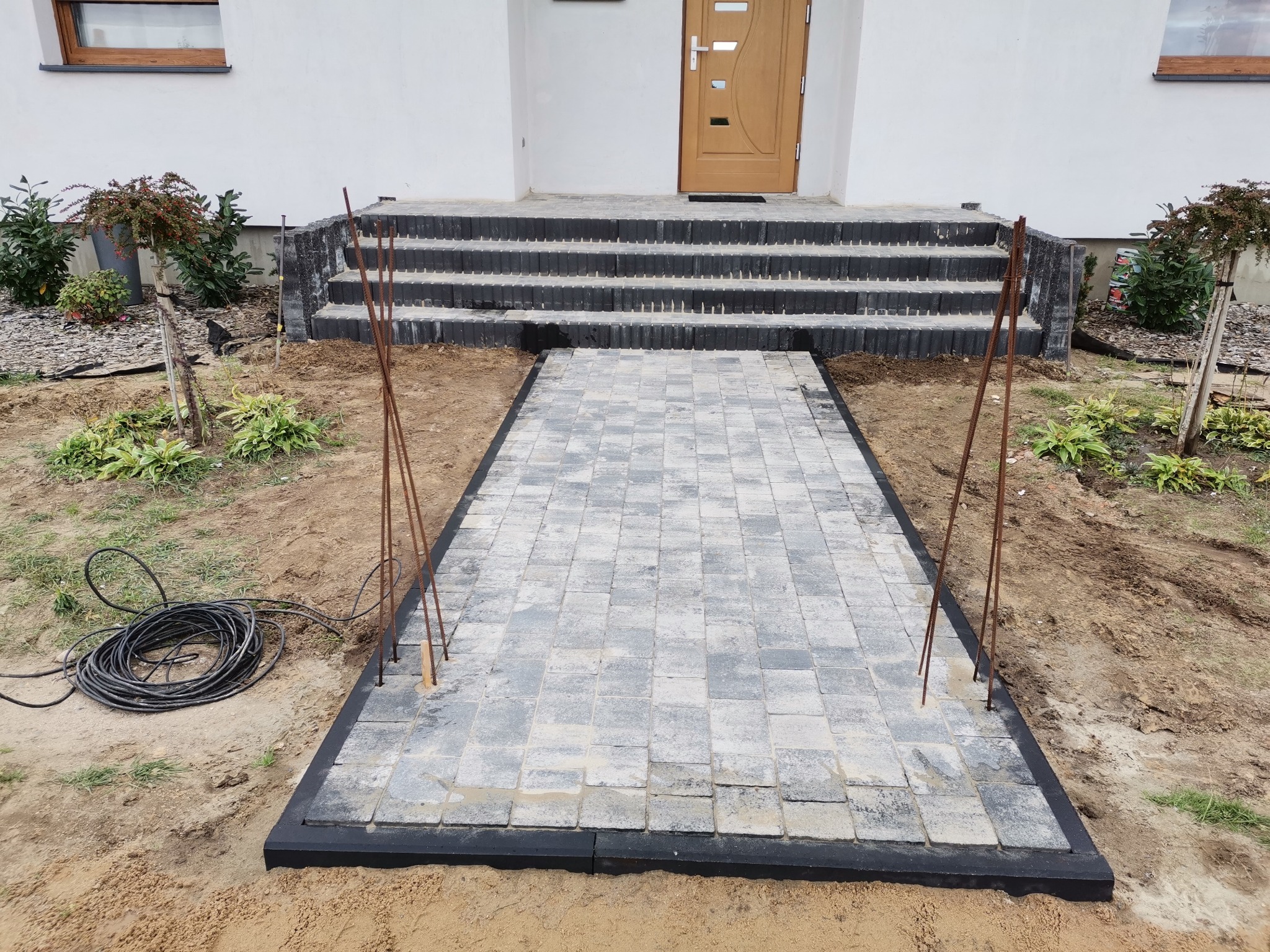 Ścieżka z szarej kostki brukowej prowadząca do schodów wejściowych domu, widoczne pręty zbrojeniowe i zwój kabla elektrycznego obok.