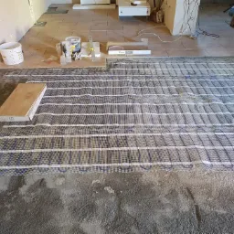 Rozłożona mata grzewcza na podłodze w trakcie montażu, widoczne narzędzia i materiały budowlane, przygotowane do dalszych prac wykończeniowych.