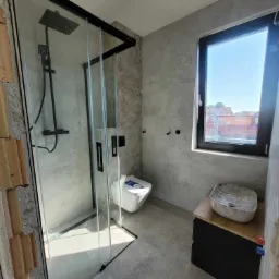 Wnętrze łazienki w trakcie wykańczania: kabina prysznicowa z czarnymi profilami, wisząca toaleta, umywalka w kształcie misy na szafce, szare płytki, widoczne niedokończone obrzeża ściany z cegłami.