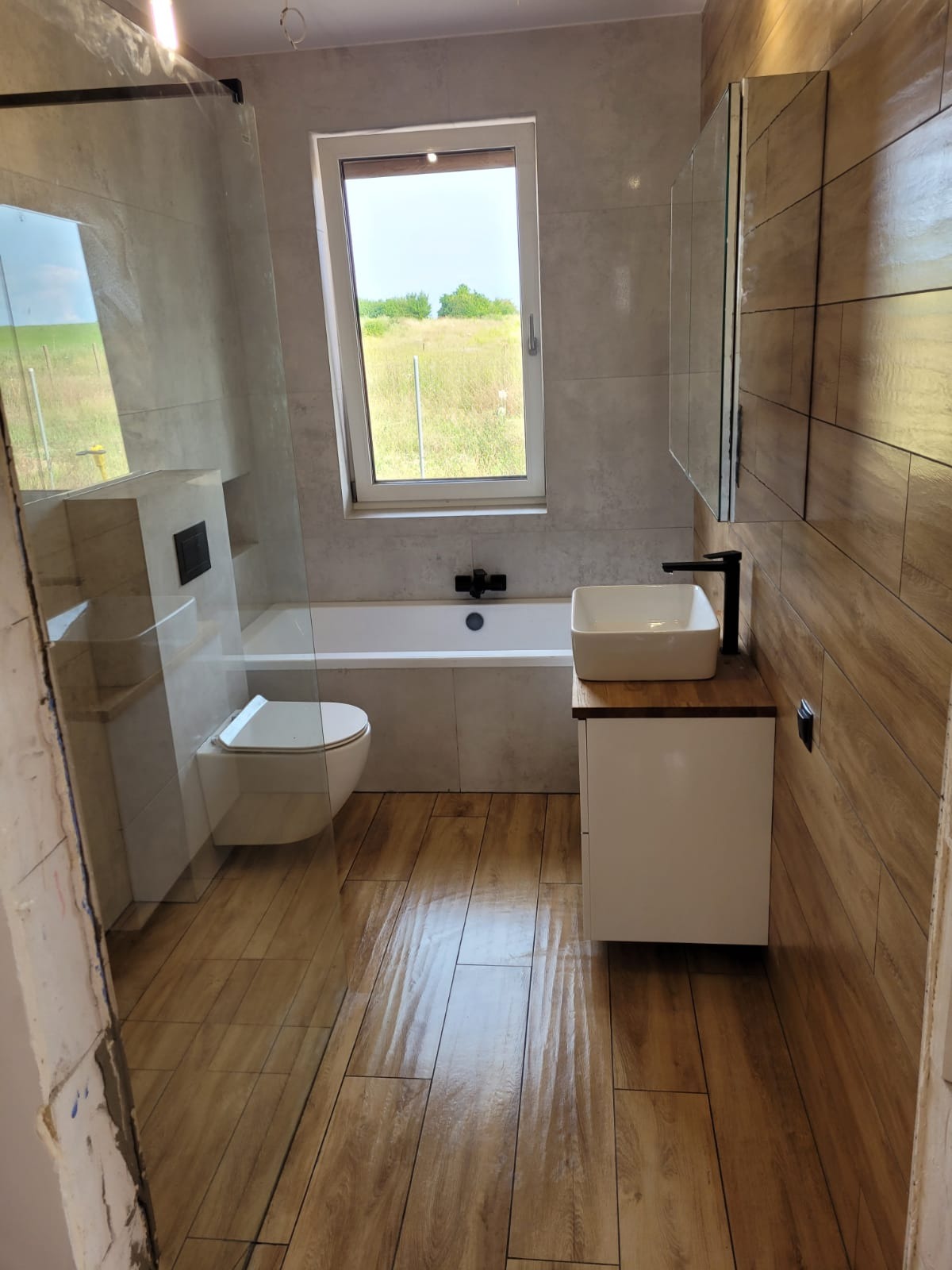 Nowoczesna łazienka z drewnopodobną podłogą i ścianą, białą armaturą i widokiem na zielone pole za oknem.