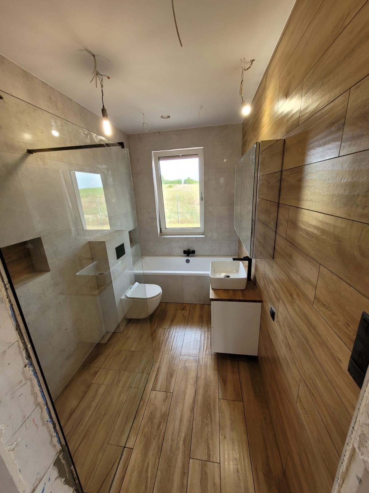 Nowoczesna łazienka po remoncie z drewnopodobnymi płytkami na podłodze i ścianie, kabina prysznicowa typu walk-in, biała wanna pod oknem i umywalka nablatowa z czarną armaturą.