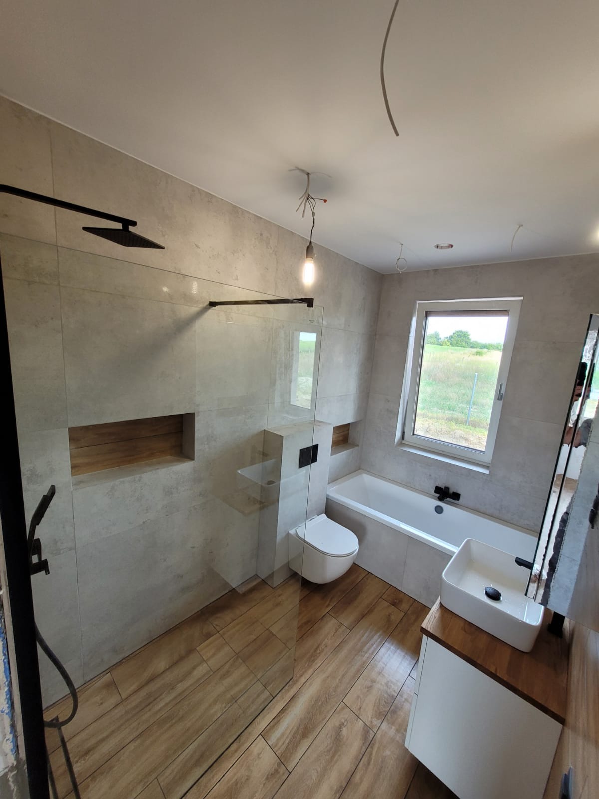 Nowoczesna łazienka z drewnopodobną podłogą, szarą glazurą, kabiną prysznicową typu walk-in z czarnymi elementami, wiszącą toaletą i umywalką nablatową na drewnianym blacie, widok na zewnątrz przez...