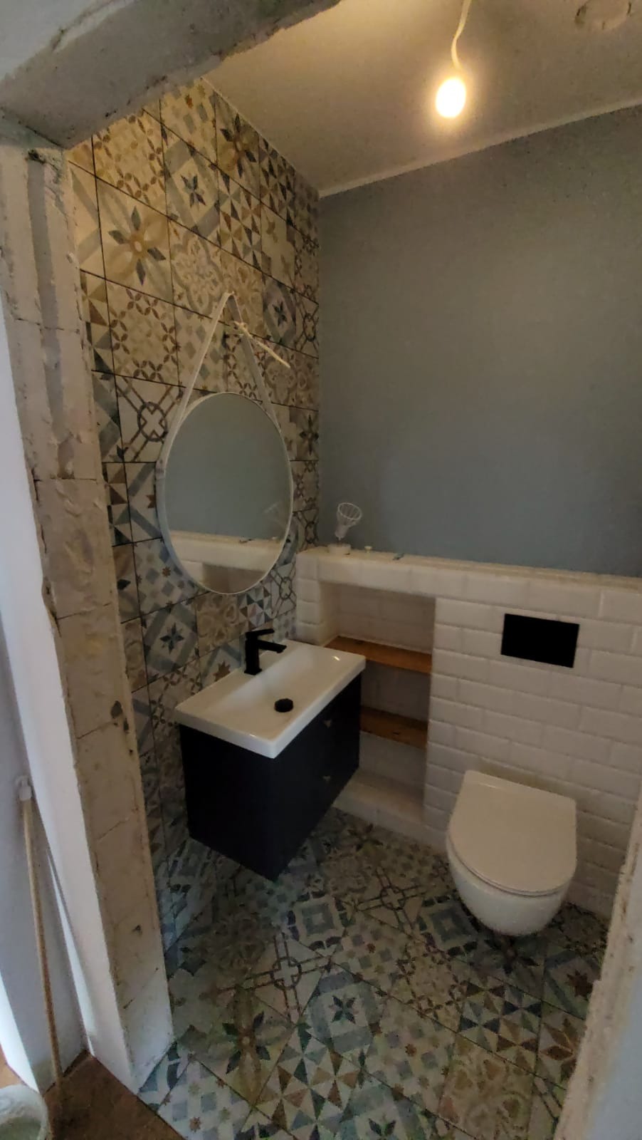 Wykończona łazienka w Rybojadach z wzorzystymi płytkami na podłodze i ścianie, umywalką z czarną armaturą, okrągłym lustrem i toaletą z zabudowaną spłuczką.
