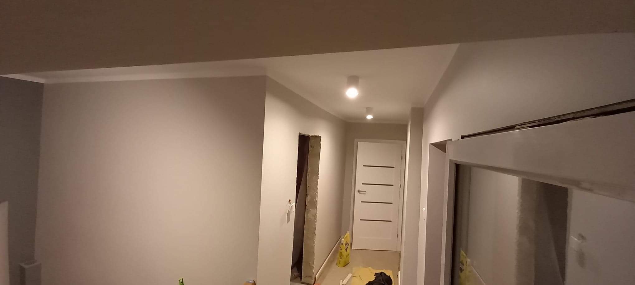 Korytarz w trakcie remontu: świeżo pomalowane ściany w odcieniach szarości z białą listwą przysufitową, widoczne przygotowania do montażu drzwi, worki z materiałami budowlanymi na podłodze.