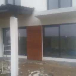 Fragment elewacji domu w trakcie prac wykończeniowych, widoczna drabina oparta o ścianę, brązowy panel elewacyjny i sterta gruzu na ziemi.