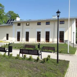 Budynek użyteczności publicznej z panelami słonecznymi na dachu, widok z zewnątrz, z ławkami i lampami parkowymi na pierwszym planie.