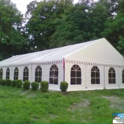 Biały namiot halowy z oknami w kształcie łuków, rozstawiony na trawiastym terenie, widoczny z zewnątrz, z zielonymi krzewami wzdłuż boku i drzewami w tle.