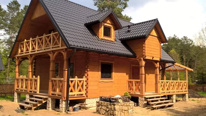 Drewniany dom z ciemnoszarą dachówką, z werandą z balustradą i kamiennym fundamentem, otoczony drzewami.