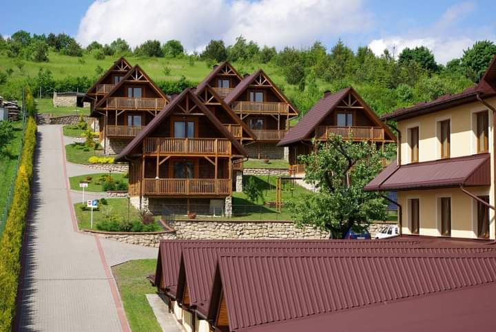 Kompleks drewnianych domków z balkonami, położonych na wzgórzu, z brukowaną drogą dojazdową i widokiem na góry w tle.