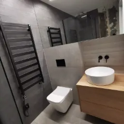 Nowoczesna łazienka z szarą glazurą, wiszącą toaletą, drewnianą szafką pod umywalką i czarnym grzejnikiem drabinkowym z podłączeniem elektrycznym.
