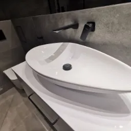 Nowoczesna, biała umywalka nablatowa o owalnym kształcie z czarną armaturą zamontowaną na szarej, betonowej ścianie. Pod umywalką biała szafka z czarnym relingiem na ręczniki. Widoczny fragment...