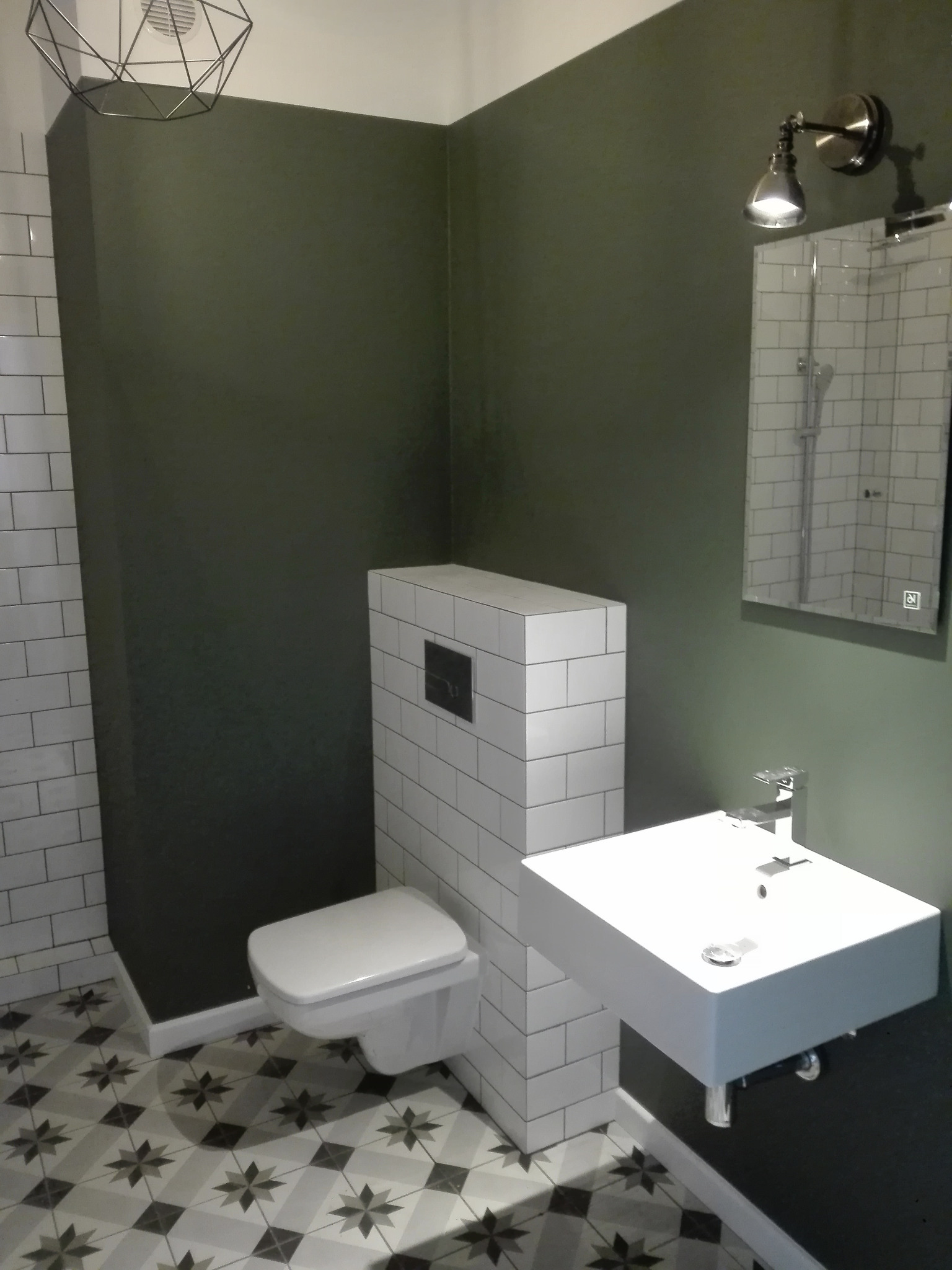 Nowoczesna łazienka z toaletą w zabudowie z białych płytek, umywalką na czarnym blacie, lustrem z oświetleniem i podłogą w geometryczne wzory.