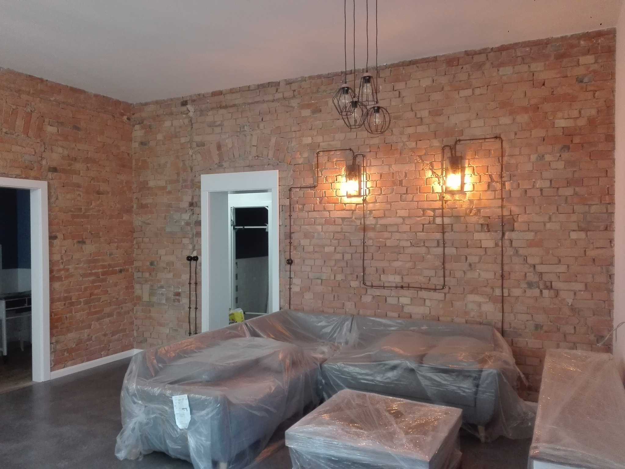 Surowe ceglane ściany we wnętrzu w trakcie remontu, meble zabezpieczone folią, widoczne instalacje elektryczne i nowoczesne lampy.