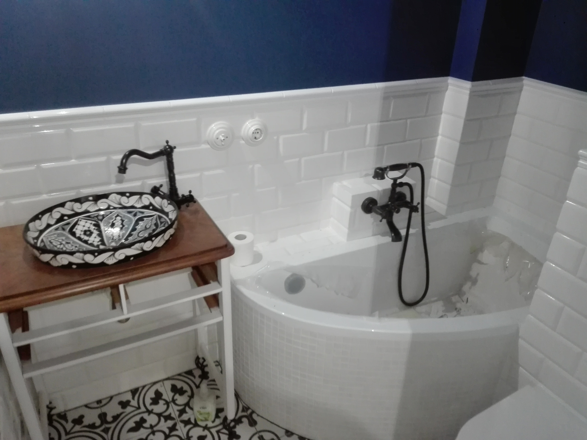 Biała łazienka z wanną obłożoną mozaiką, umywalką na drewnianym stelażu, czarną armaturą i dekoracyjną posadzką w czarno-białe wzory.